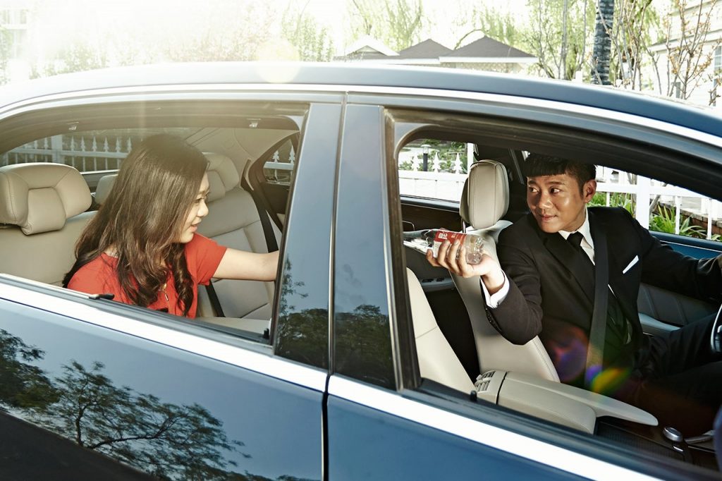 A driver in a black suit offers a bottle of water to a female passenger in the backseat