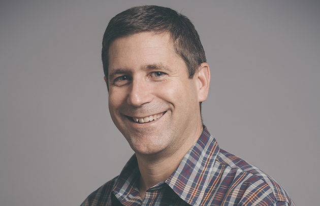 Headshot portrait of a man smiling, wearing a plaid shirt
