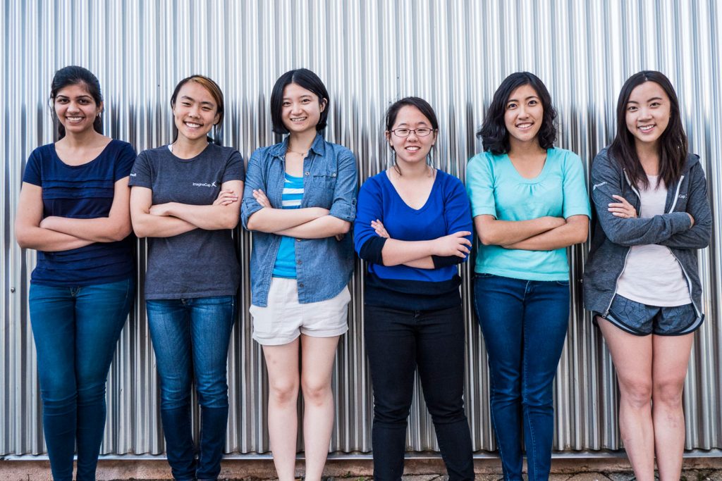 6 young women pose, arms crossed, standing in a row against a corrugated steel wall
