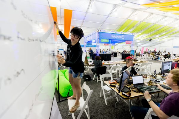 A past Hackathon participant writing on a whiteboard in a room full of people working on various projects at tables