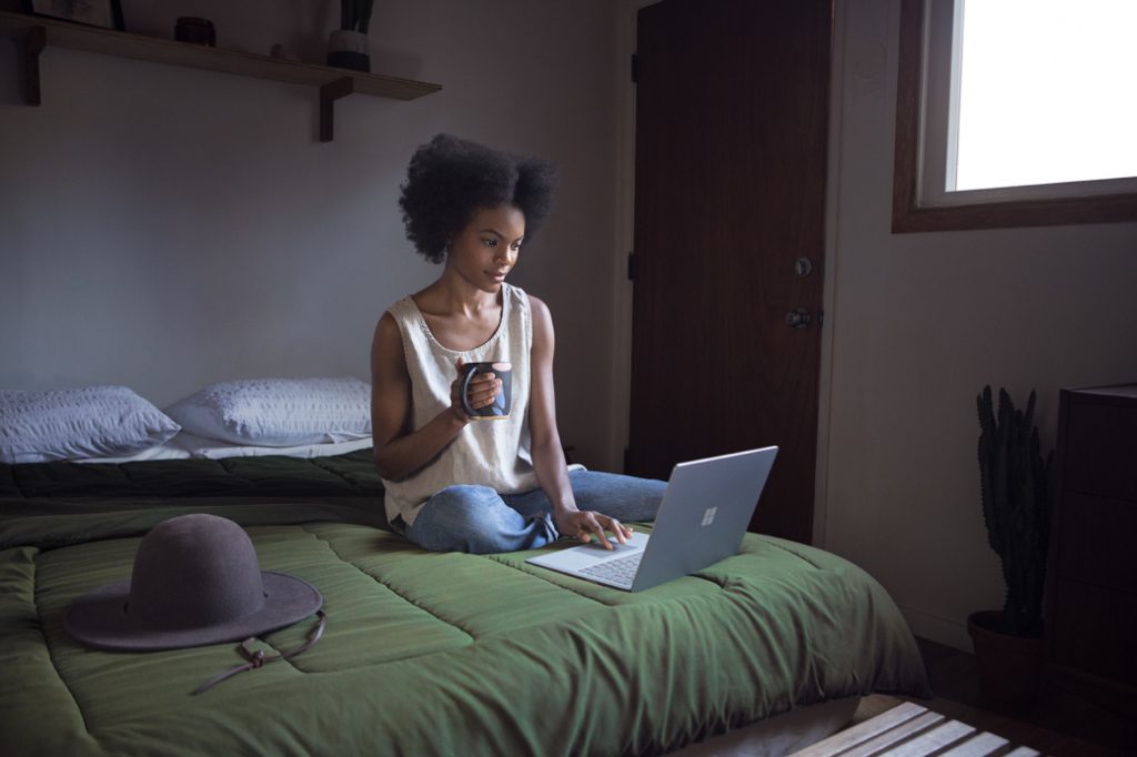 Woman sitting on bed using Surface with cup of coffee in one hand