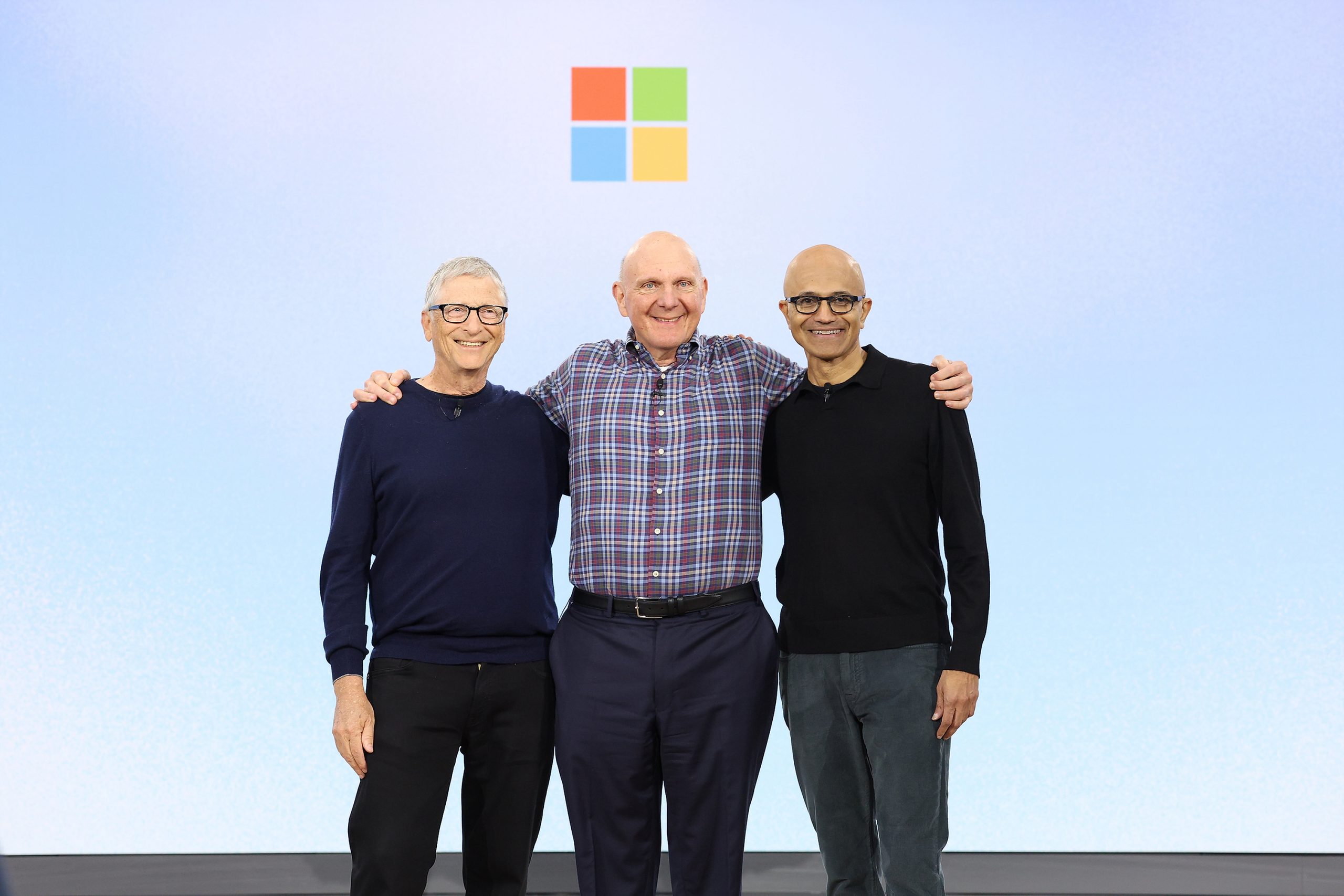 Three men with their arms around each other’s shoulders stand on stage in front of the colorful square Microsoft logo.