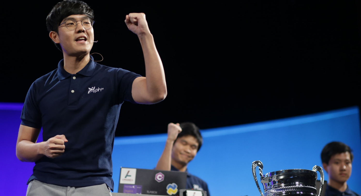 Photo of three Imagine Cup competitors on stage, two with a fist raised as if celebrating, with a trophy in the foreground