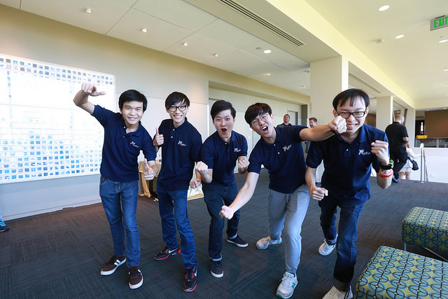 Photo of five enthusiastic young men wearing matching navy shirts pose and grin