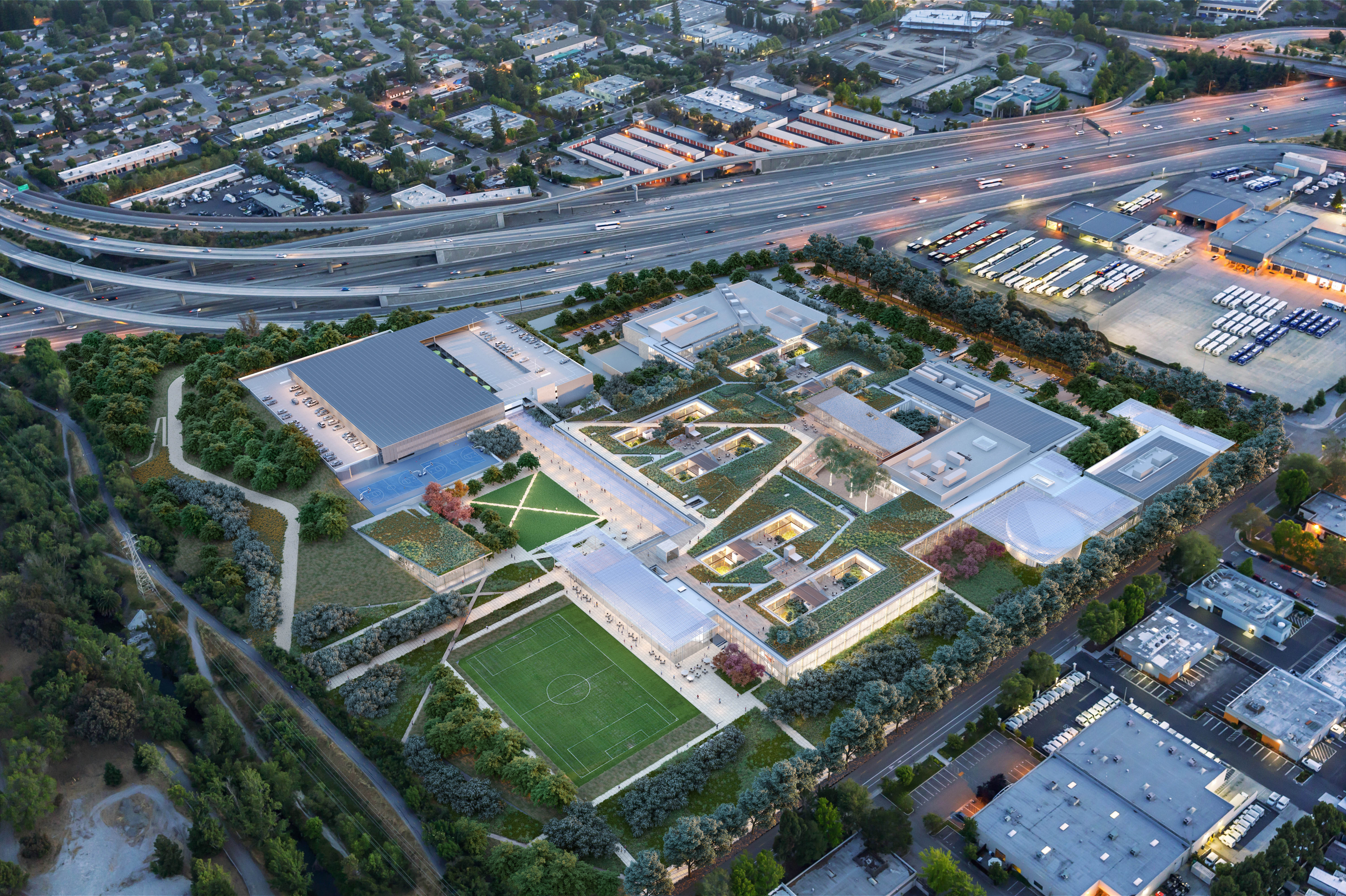 Photo shows aerial view of sleek buildings and a soccer field surrounded by greenery next to a freeway
