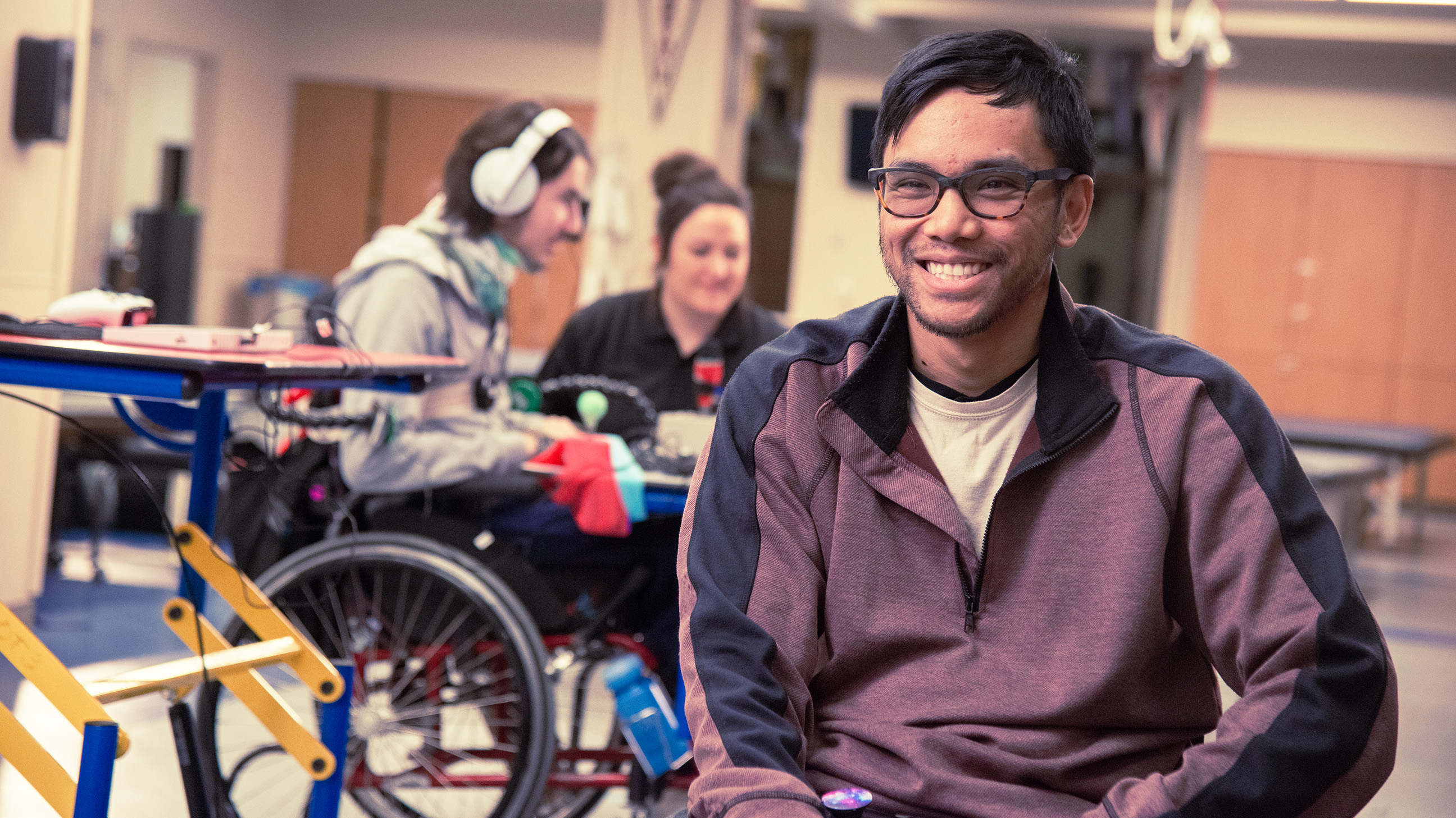 Photo of a man smiling in the foreground and a woman in the background who is working with a man in a wheelchair and using headphones