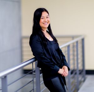 Photo of Lili Cheng leaning against railing inside a modern office building and smiling
