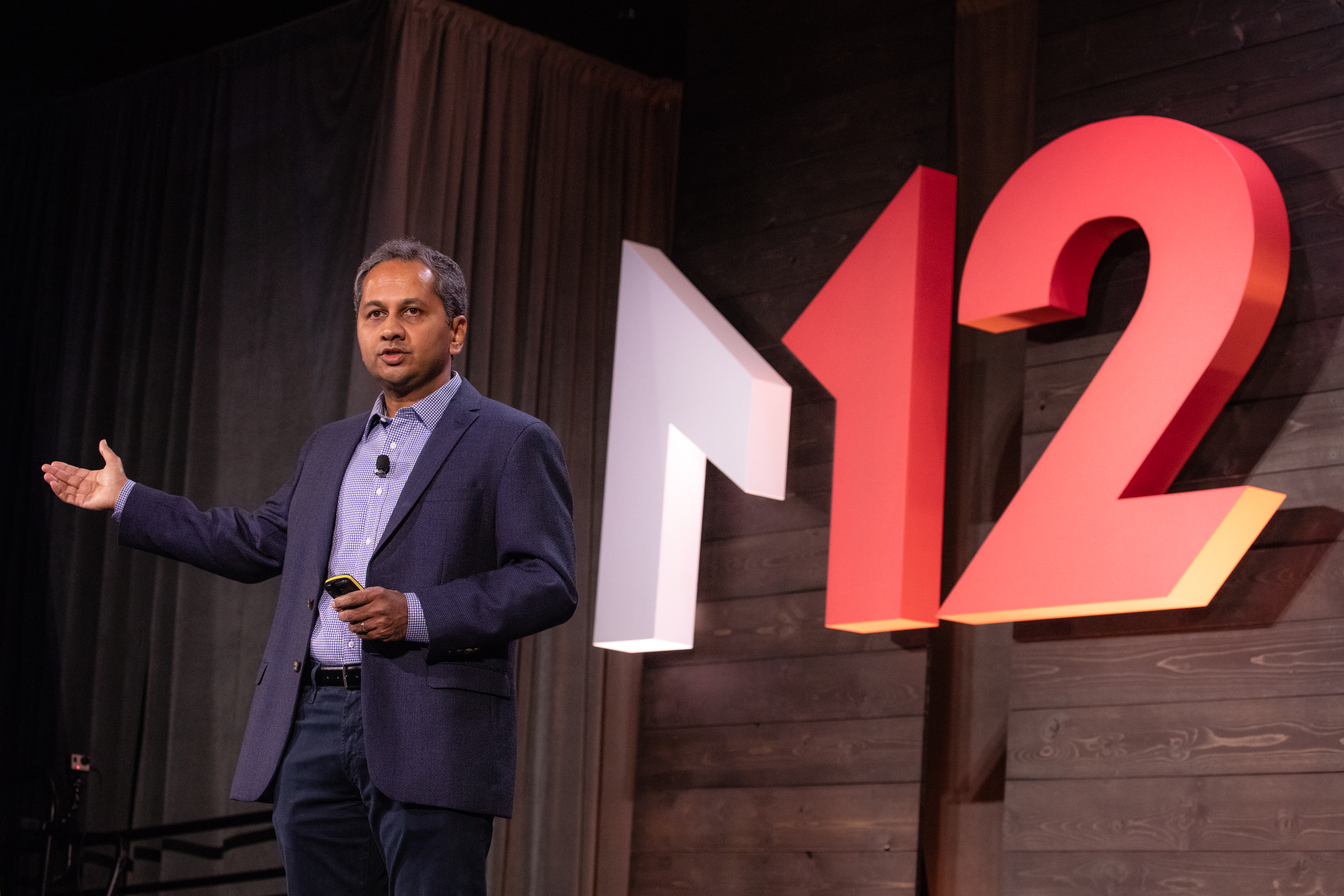 Nagraj Kashyap standing onstage at the M12 Summit in front of large M12 sign.