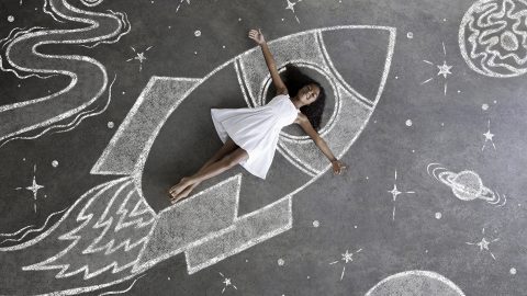 Barefoot girl in white dress explores a whimsical chalkboard universe inside her chalkboard spaceship, arms wide open