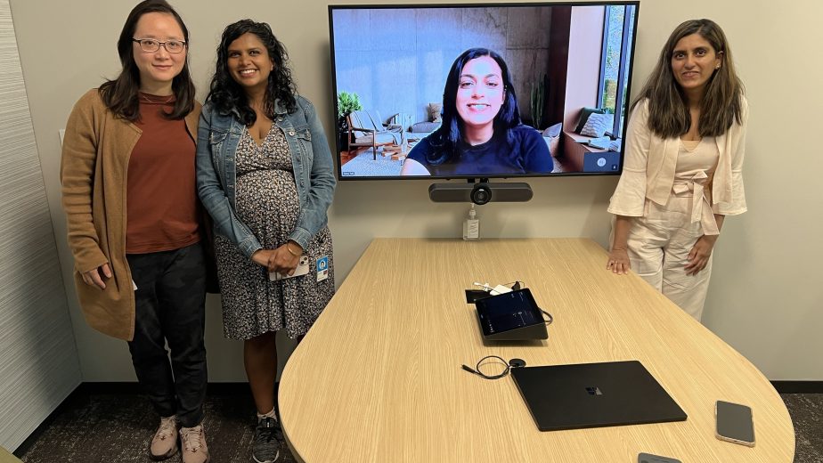 Three women in a conference room and one on a screen.