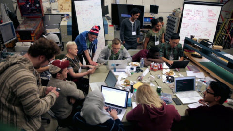 Individuals work on laptops around a large table, with whiteboard drawings in the background.