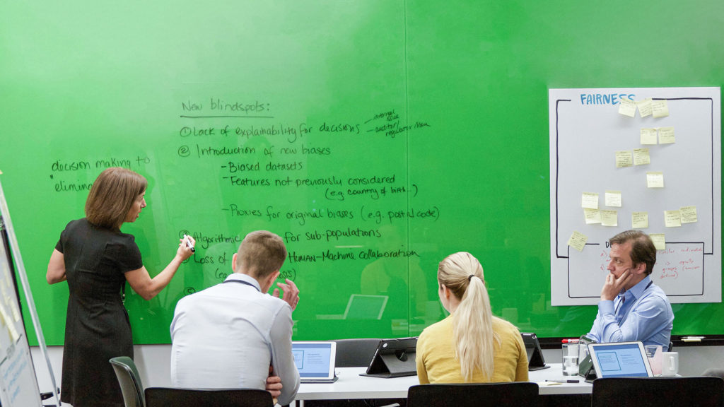 People sit in front of a white board. Written on the whiteboard are words inculding fairness, blindspots and explainability