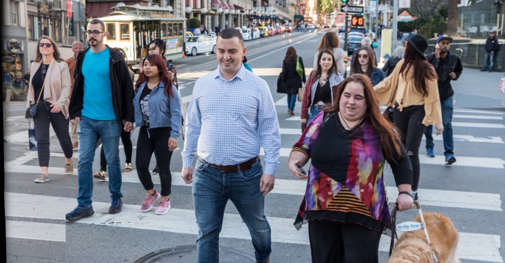 Image of Alex and Maia crossing a busy intersection in downtown San Francisco. Maia is using Soundscape which has given her information about the layout of the intersection in 3D audio