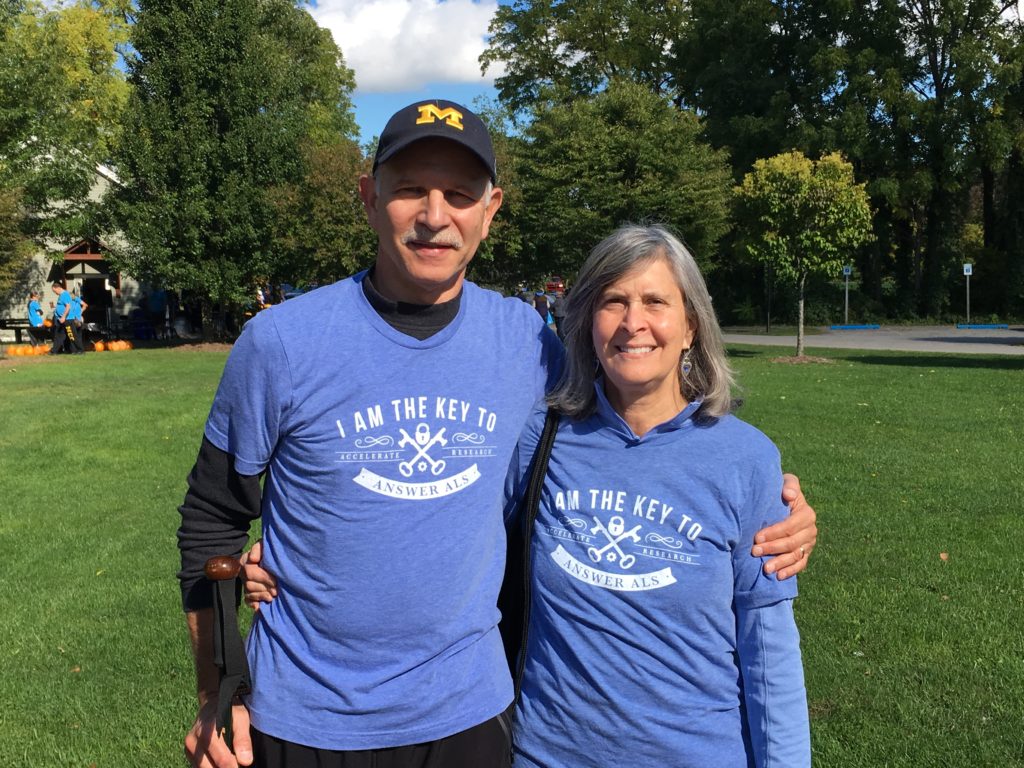Joel Shamanski and his wife Ann, stand next to each other, outside, at the Answer ALS Walk in Rochester New York. They are both wearing Answer ALS shirts.