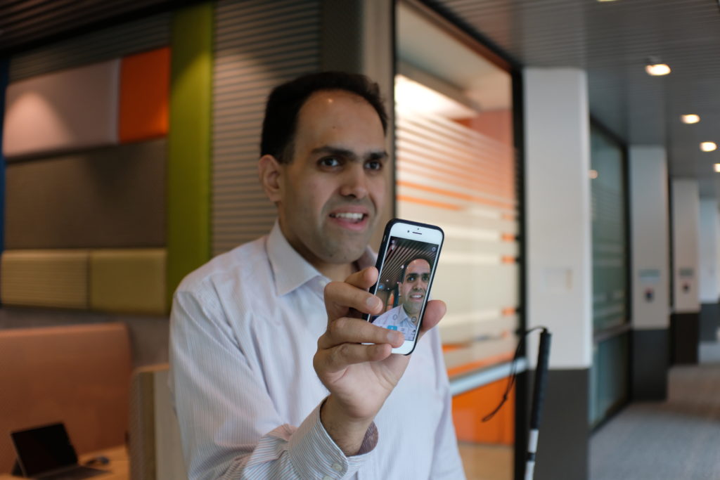 A business man in an office setting holding up a smartphone that shows an image of himself.