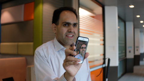 A business man in an office setting holding up a smartphone that shows an image of himself.