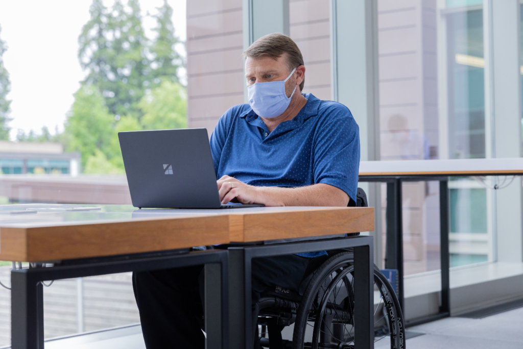 A male employee in a wheelchair working on his Surface laptop at a desk wearing a face mask.