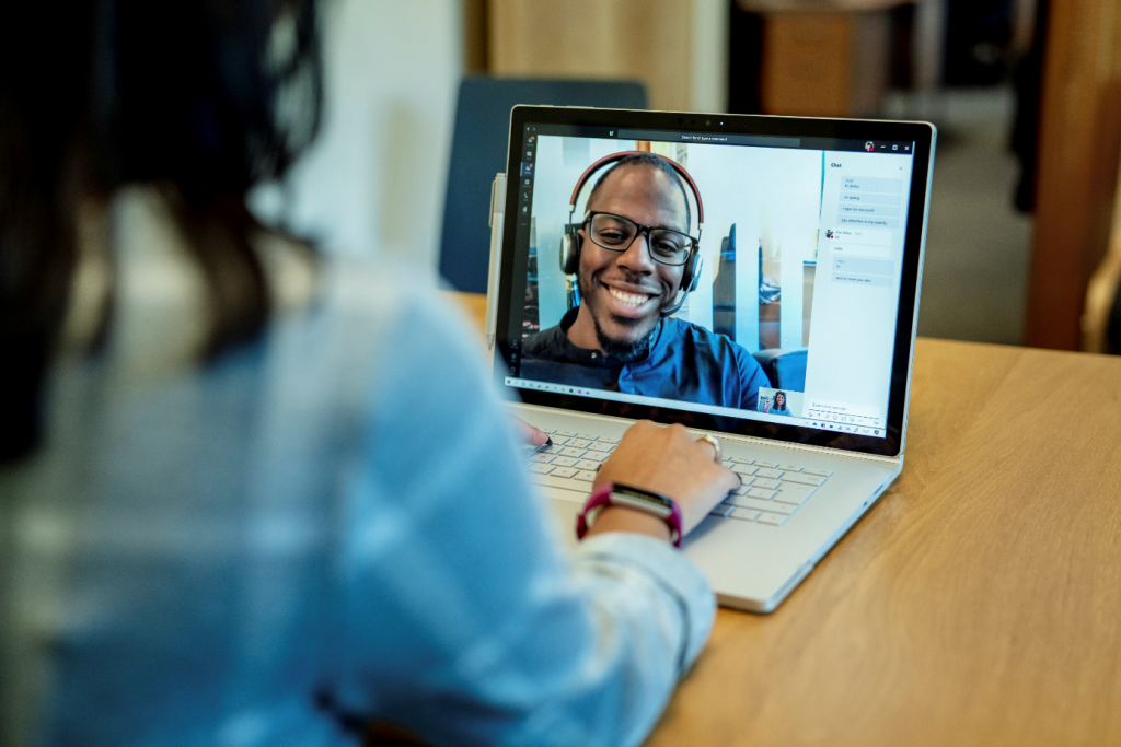 Woman at a desk using a Surface laptop to make a Microsoft Teams video call with one man smiling and wearing a headset.