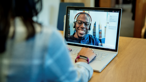 Woman at a desk using a Surface laptop to make a Microsoft Teams video call with one man smiling and wearing a headset.