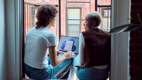 Two Gen Z friends sitting on fire escape balcony with backs to camera laptop open with Windows 11 start screen and start menu. Keywords: young man; young woman; inside; sitting; window; sill; interior; casual; city; balcony; New York City
