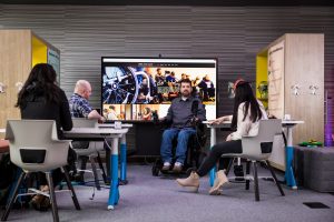 A group of people sitting at desks and one person in a wheelchair discuss design in the inclusive tech lab.