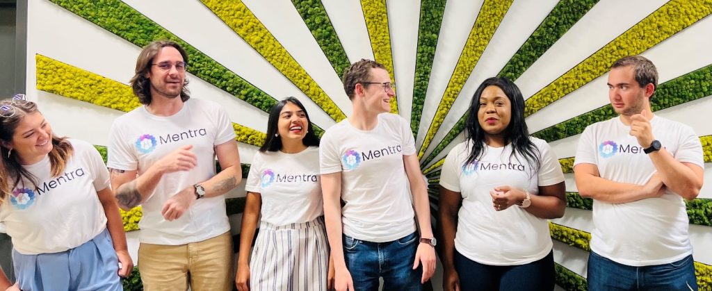 7 of the Mentra employees wearing branded Mentra t-shirts against a background with colorful yellow and green strips.