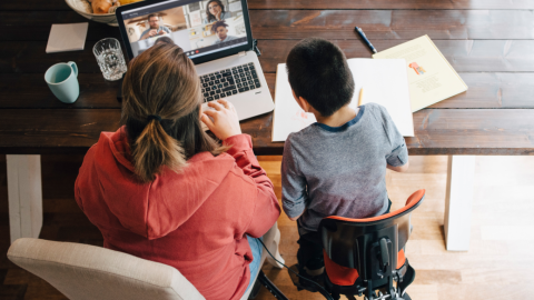 A mother working from home, in a Microsoft Teams meeting, while her son is seating next to her.