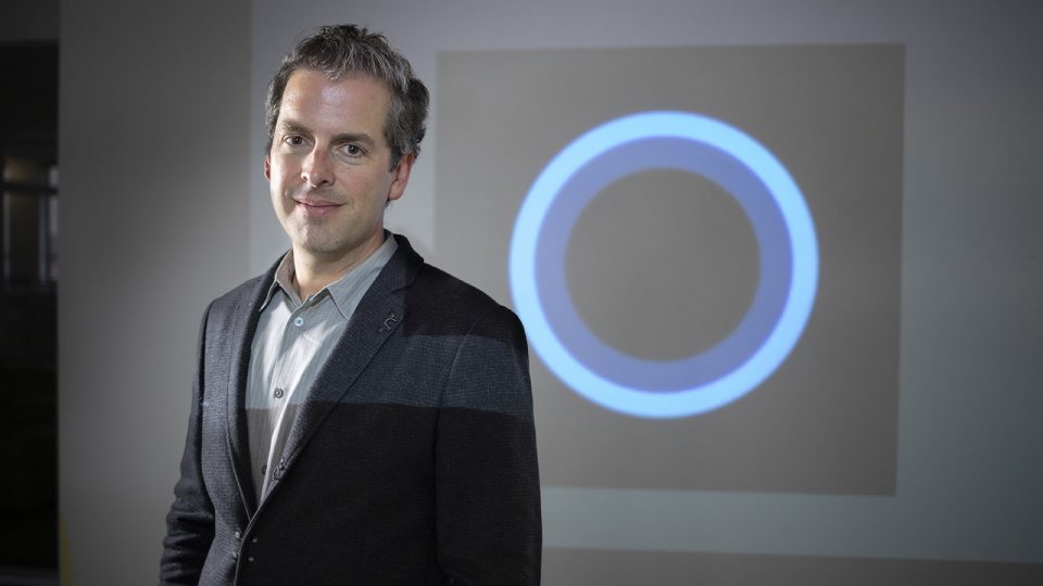 Javier Soltero stands in front of a projected image of the Cortana logo over his left shoulder, looking at the camera, smiling