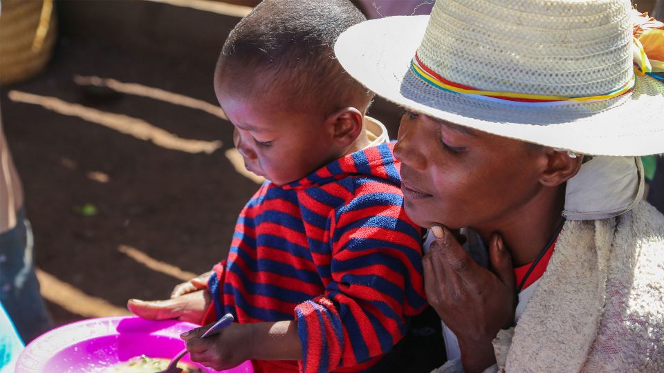 A mother watches the toddler sitting on her lap eating soup from a pink plastic bowl