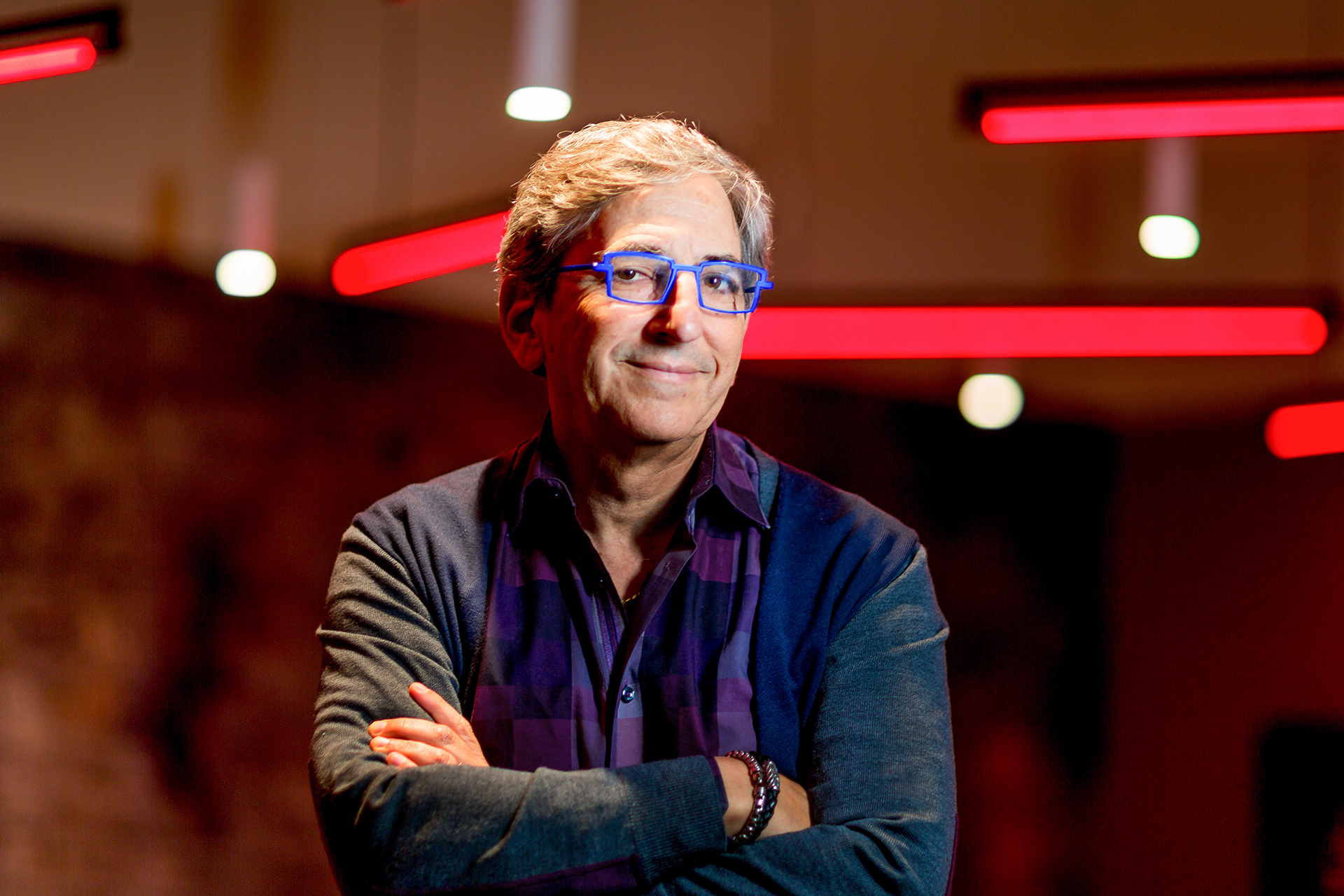 Norm Judah looks into the camera with arms folded, standing in front of red neon lighting