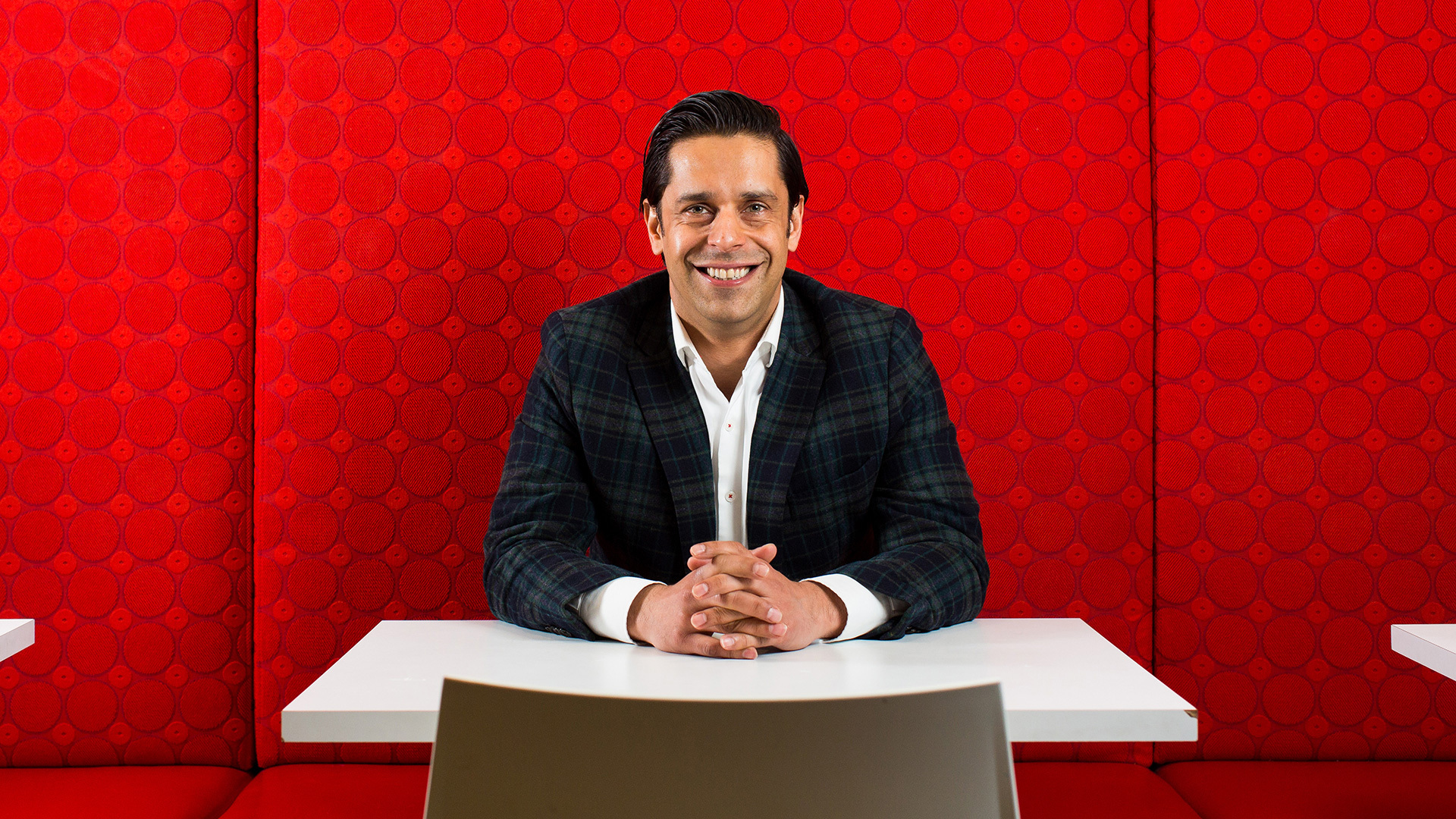 Bharat Sandhu sitting at a table with arms folded, sitting in front a bright red background