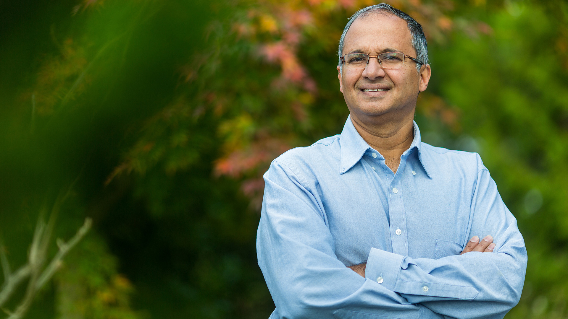 Arul Menezes standing with arms folded in front of green foliage in the background