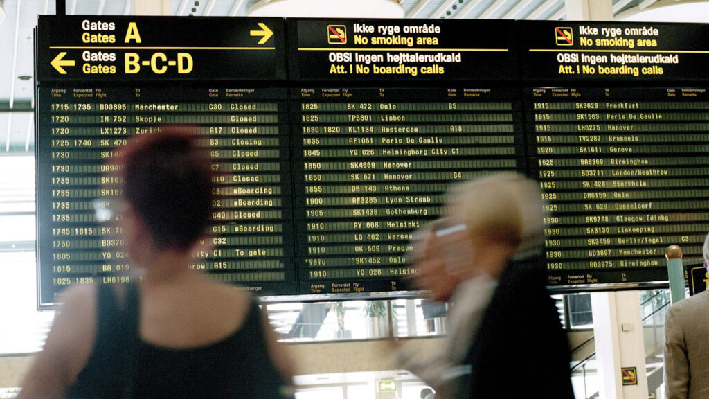 A busy computer terminal with blurry people in the foreground and an arrival/departures board in the background