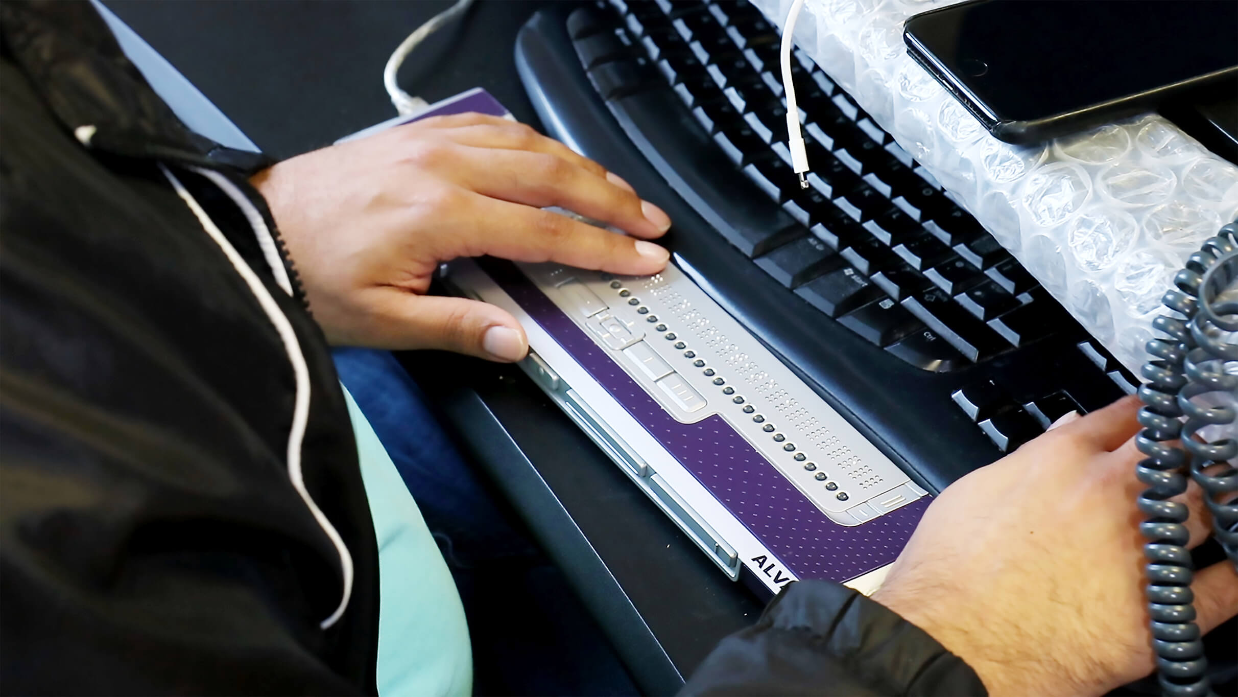 Photo of a man’s hands on a braille display next to a lettered keyboard