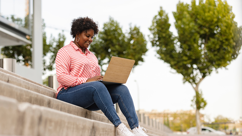 Photo of woman sitting on a step outside with a laptop on her lap.