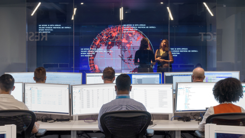 Conference room with people sitting in front computer screens while two women are seen standing in front of a digital globe with red markers over different areas.