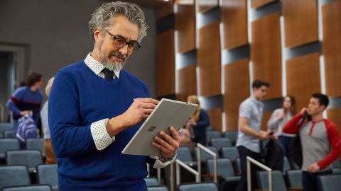 A male university instructor in a lecture hall, using Surface Go to prepare course materials while students file in.